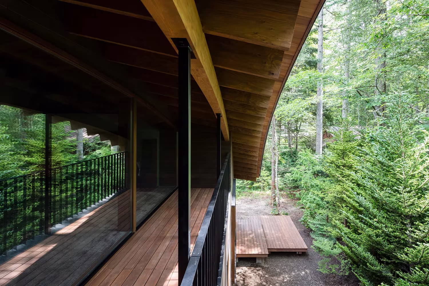 Galería con barandilla metálica y alero curvo de madera abierta al bosque en la Casa en Karuizawa