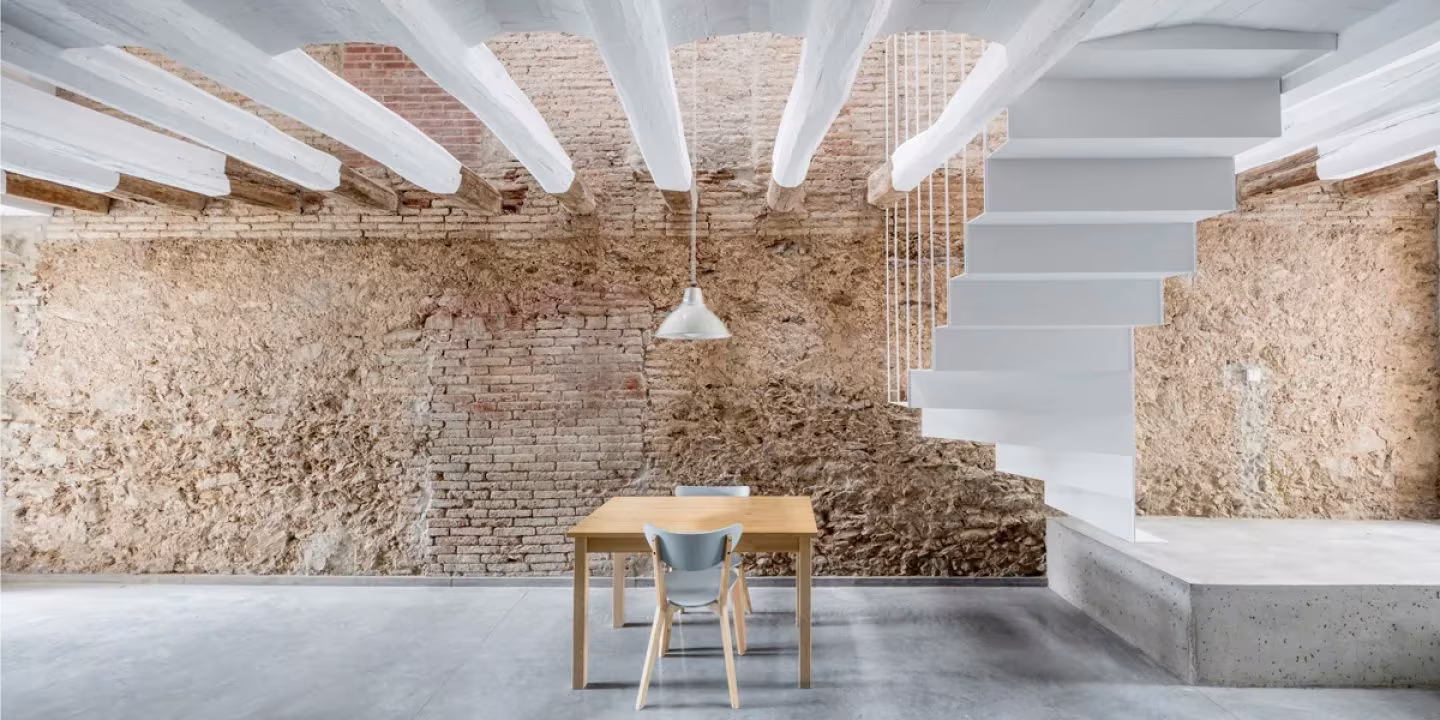 Planta baja con mesa de madera, vigas blancas y muro de ladrillo visto en la reforma de DataAE, Sant Feliu