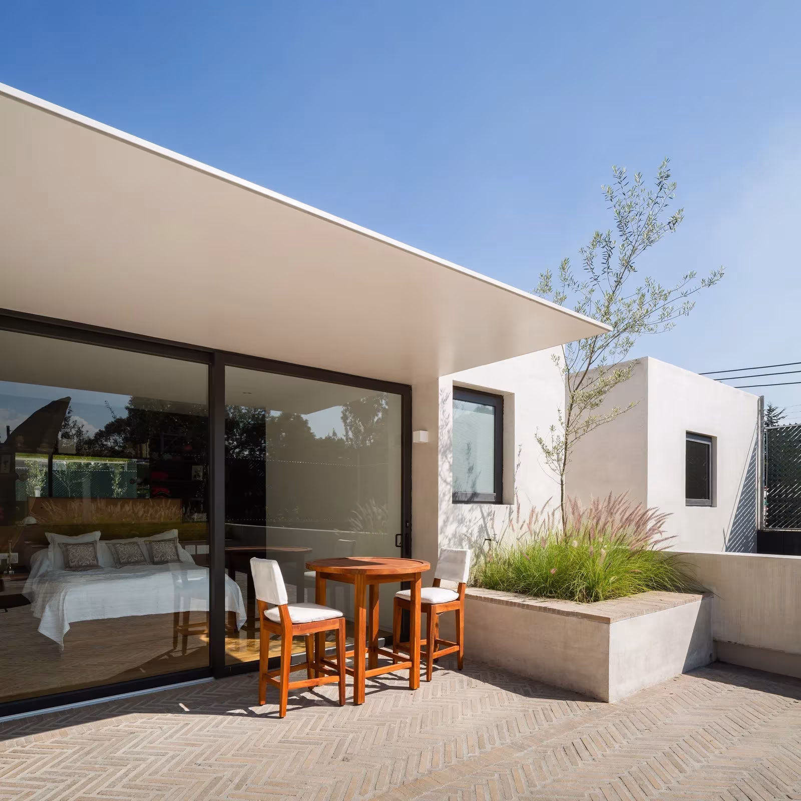 Terraza con mesa y sillas de madera ante el dormitorio acristalado de la Casa Jardín 58 de DCPP Arquitectos