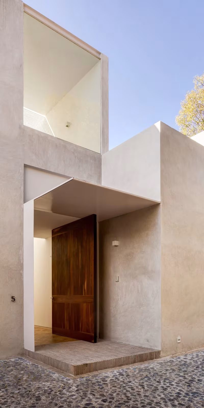 Acceso a la Casa Jardín 58 con puerta de madera pivotante y torre blanca de hormigón con lucernario