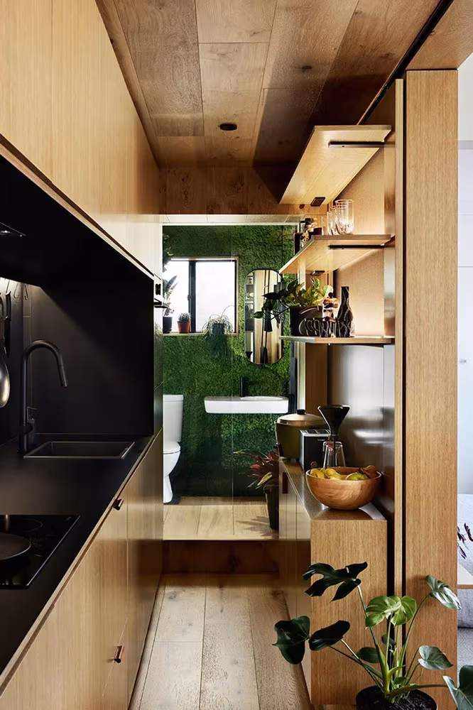 Pasillo de roble con cocina negra y baño con pared vegetal al fondo en el apartamento de Jack Chen