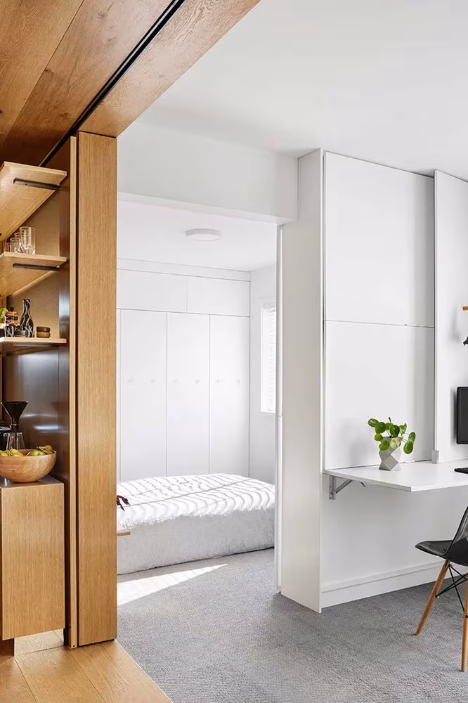 Cama blanca plegada y escritorio abatible vistos desde el pasillo de roble del apartamento de Jack Chen
