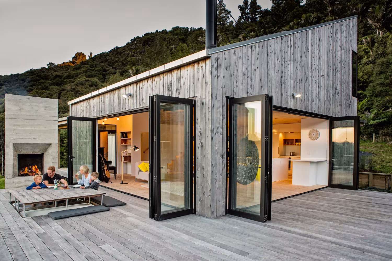 Back Country House de LTD con las puertas plegables abiertas a la terraza y la chimenea de hormigón al fondo