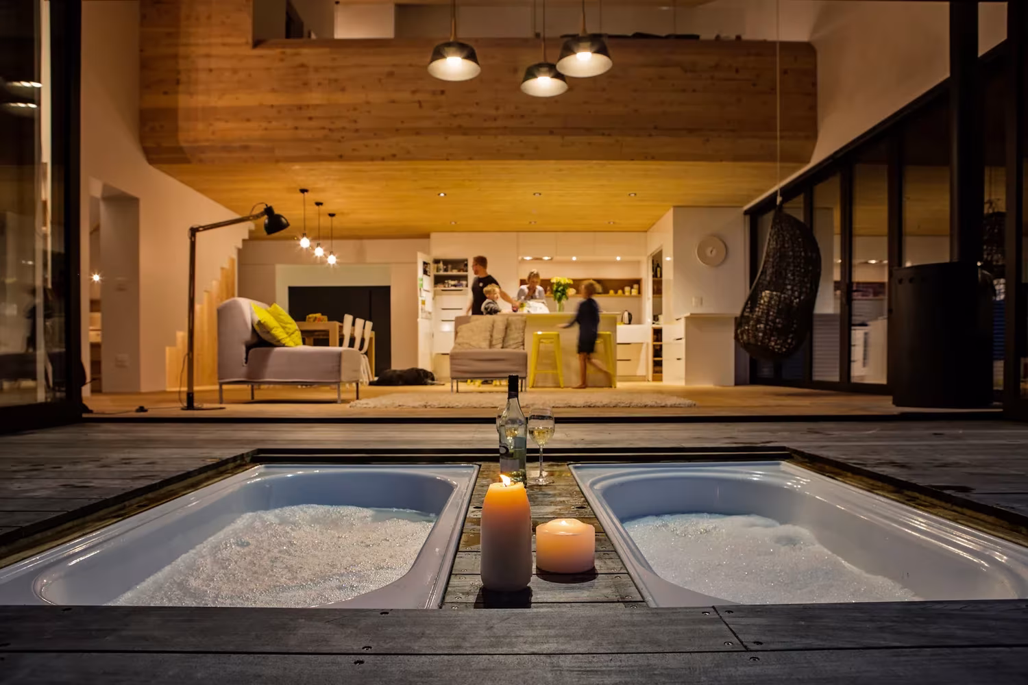 Bañeras empotradas en la terraza de madera con la sala iluminada al fondo en la Back Country House, Puhoi