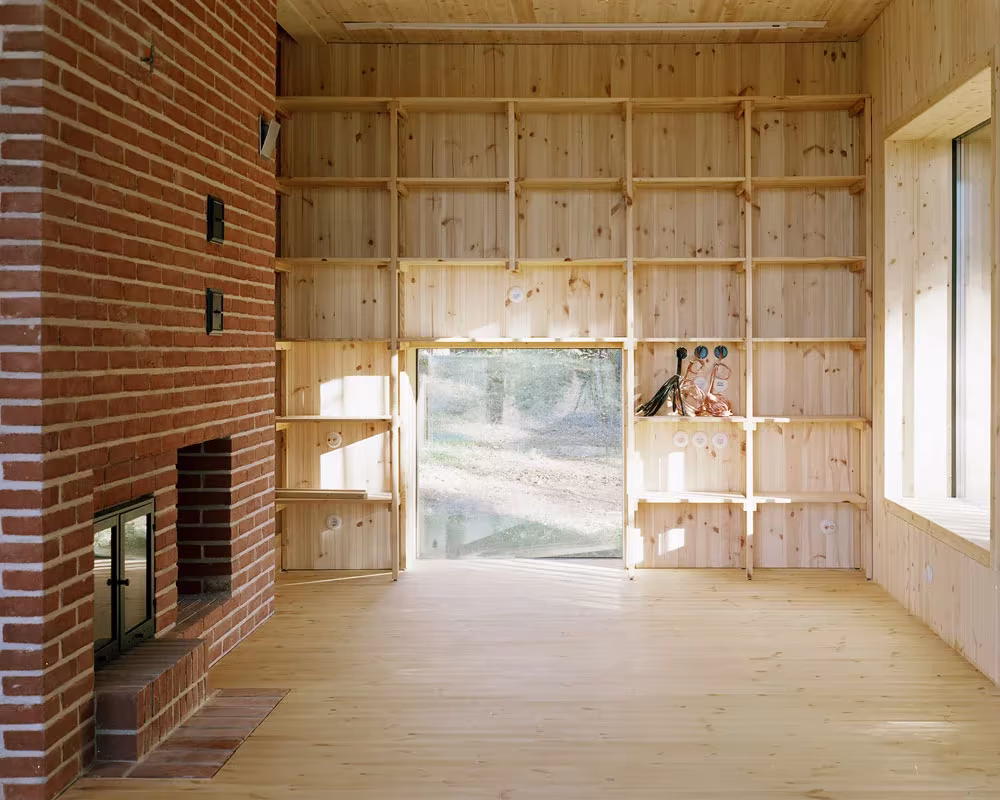 Sala de pino con chimenea de ladrillo, estantería a la altura del techo y ventana al bosque en la Villa Idun-Lee