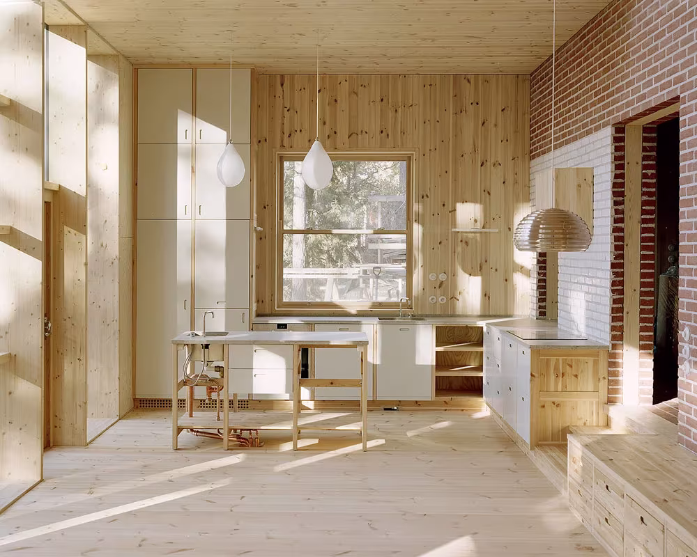 Sala con cocina blanca, mesa de madera y muro de ladrillo bajo la luz del bosque en la Villa Idun-Lee
