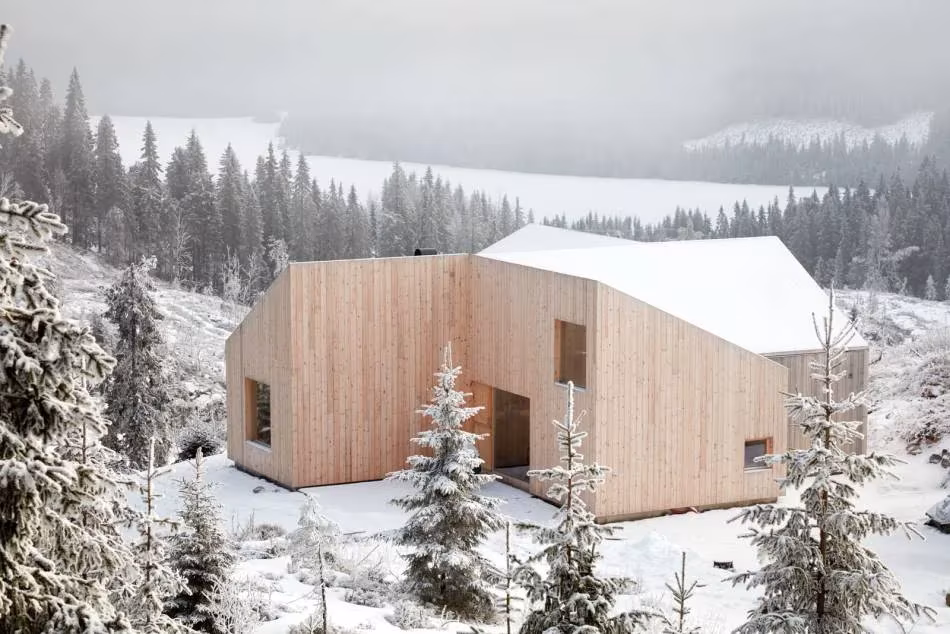 Cabaña de madera clara con cubierta quebrada y nieve sobre el lago Mylla, de Mork Ulnes Architects