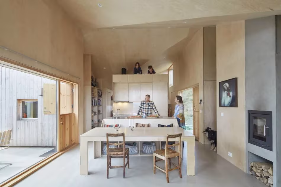 Comedor con mesa de madera, cocina y estufa de leña bajo techo de madera clara en la Mylla Hytte, Noruega