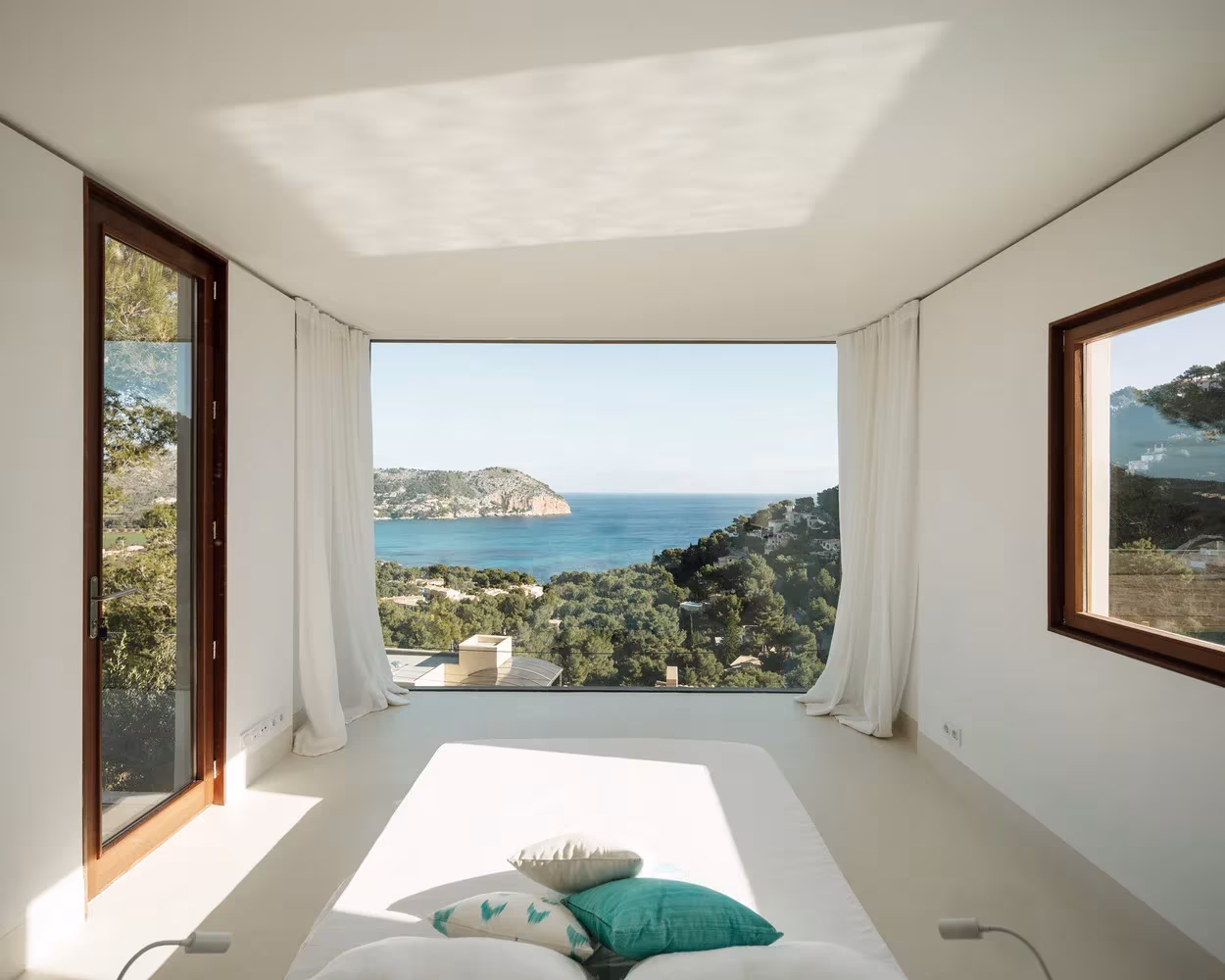 Dormitorio de la Casa Can Canyís con ventanal enmarcado sin cristalera que abre al mar y cortinas blancas