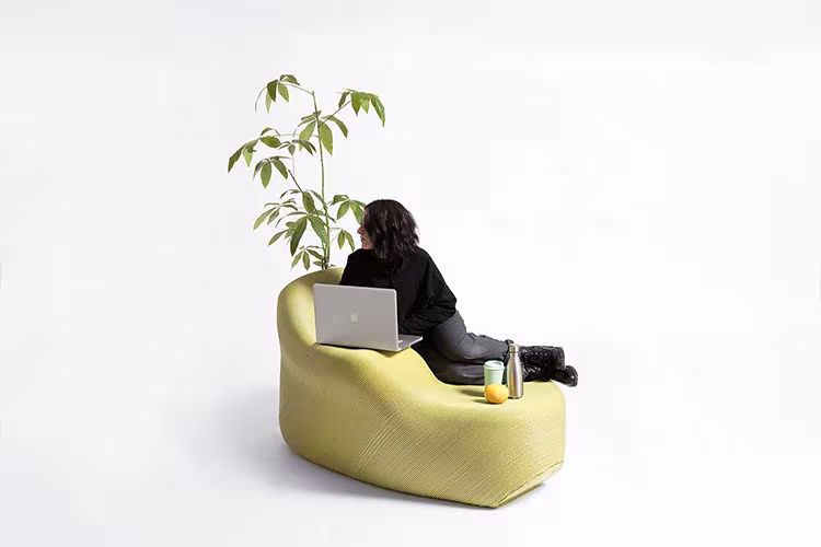 Banco amarillo de plástico reciclado de The New Raw con una maceta y una persona sentada con un portátil