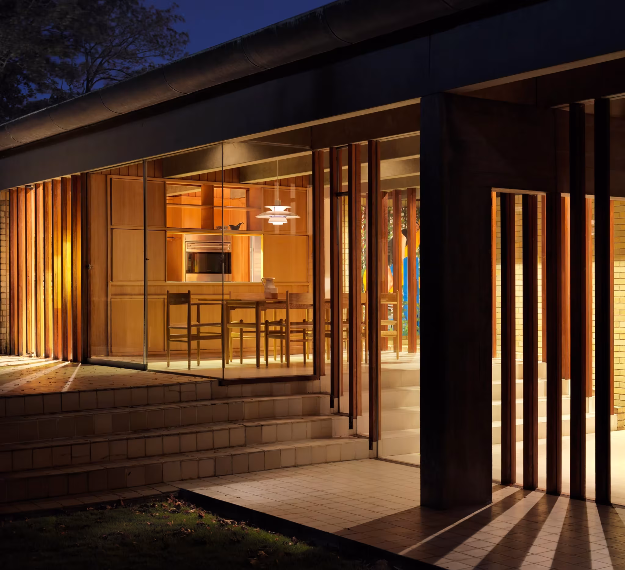 Fachada acristalada de la Ahm House de Jørn Utzon iluminada al anochecer tras pantallas verticales de madera