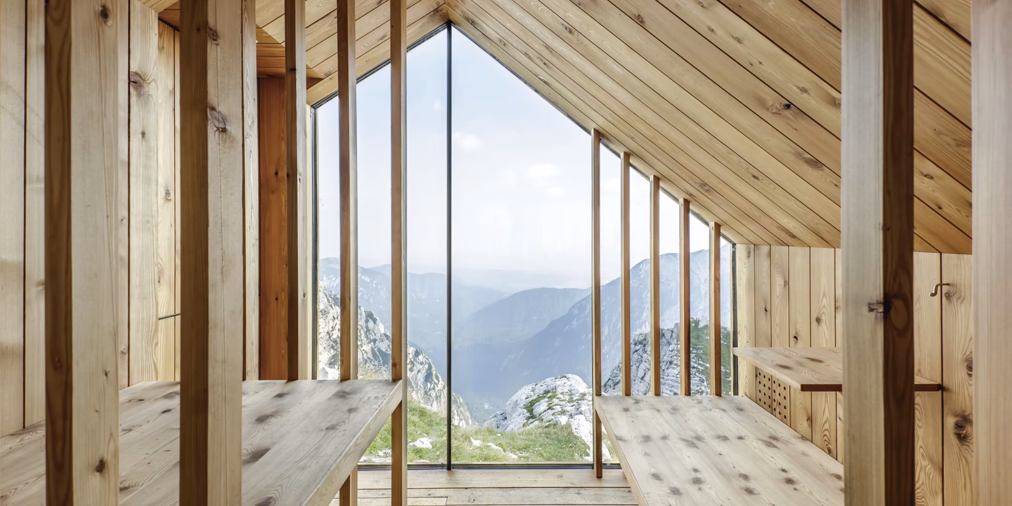 Interior de madera del refugio Skuta con ventanal de suelo a techo y mesas sobre el valle de los Alpes eslovenos