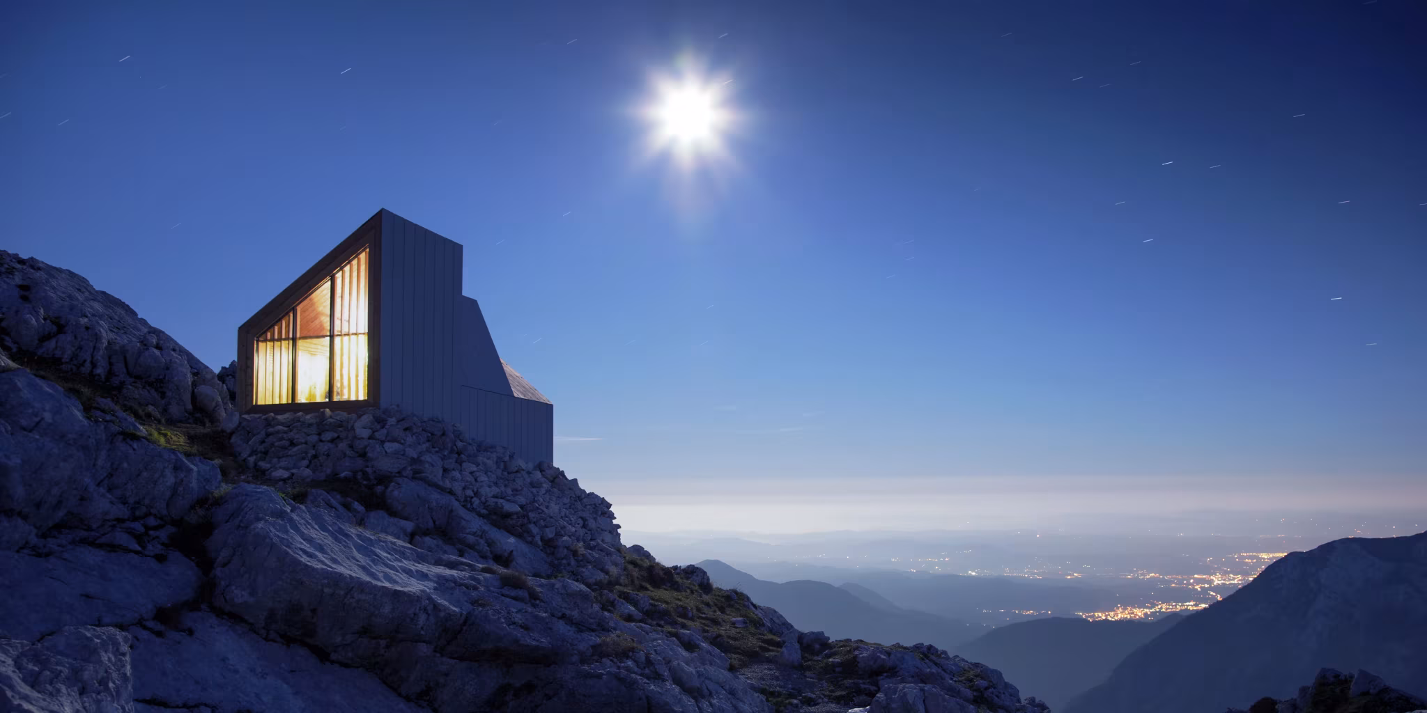 Refugio Skuta de OFIS Arhitekti con su ventana iluminada bajo un cielo nocturno sobre el valle, Alpes eslovenos