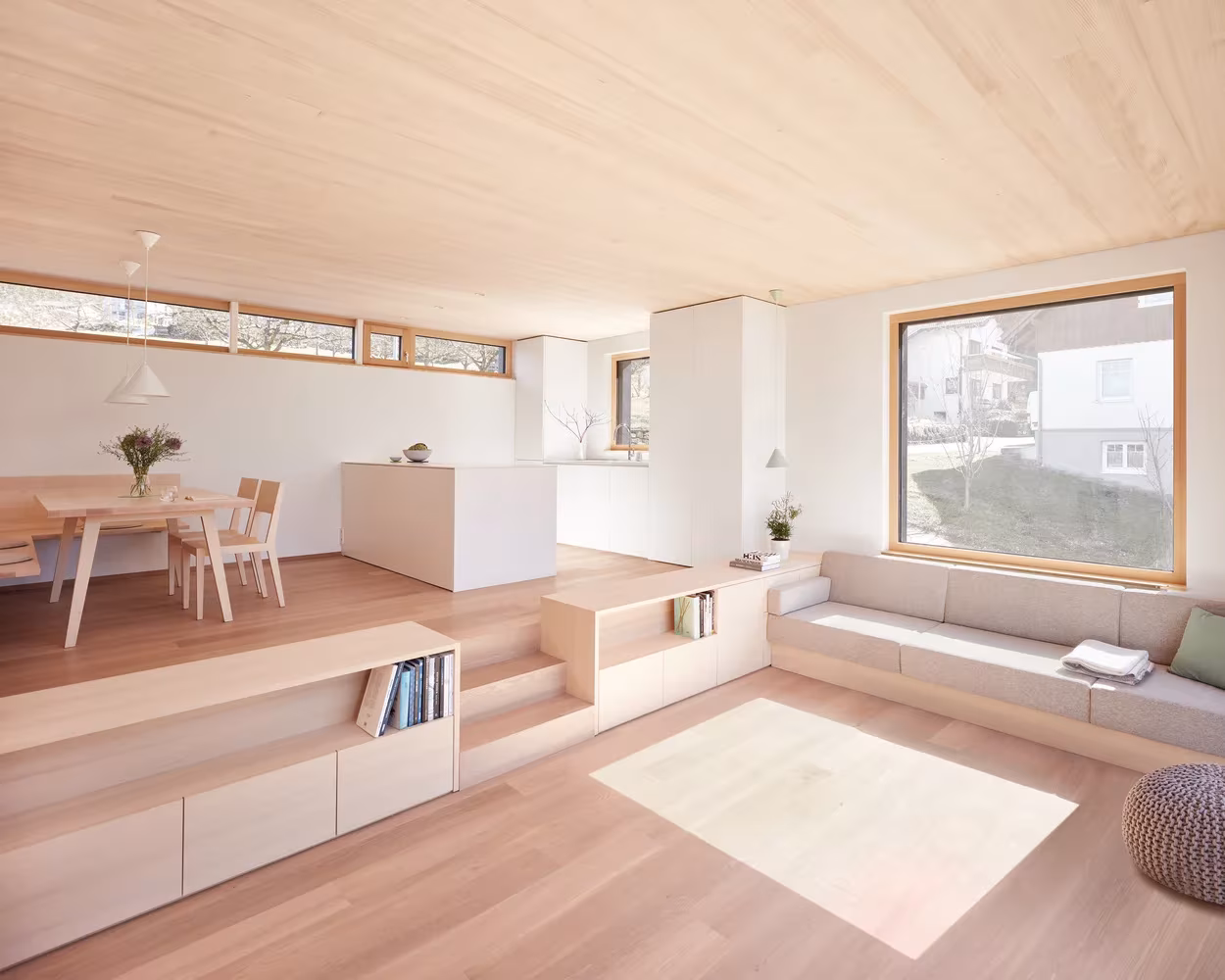 Salón con zona de estar elevada, banco corrido y ventana de marco de madera en la casa de MWArchitekten, Hohenems