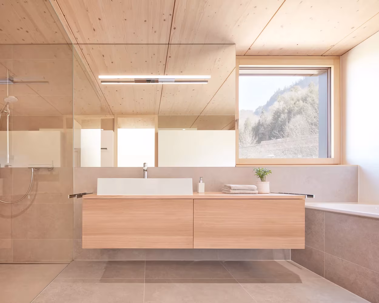 Baño de madera clara con mueble suspendido, ducha de vidrio y ventana al paisaje en la casa de Hohenems, Austria