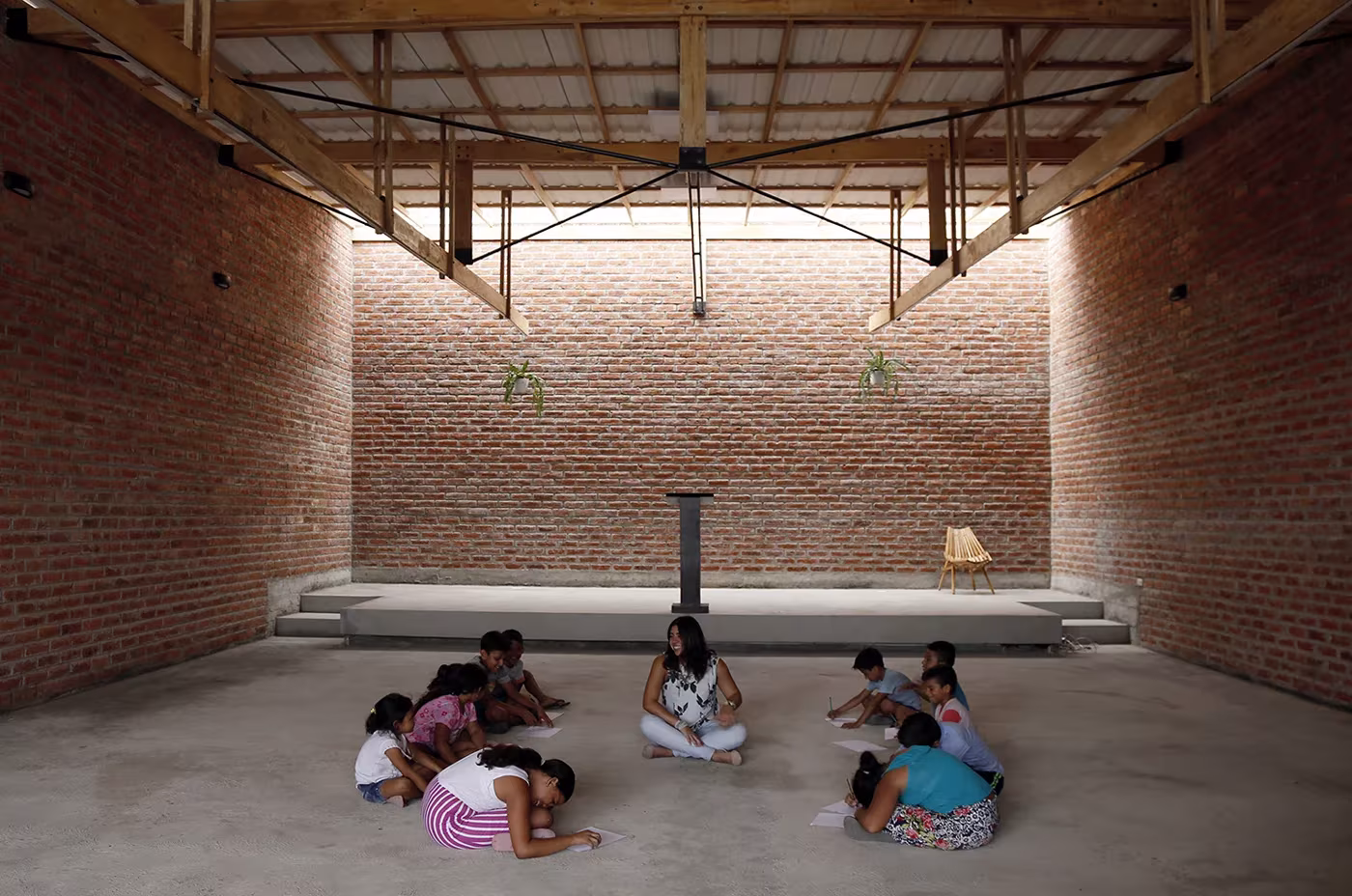 Niños sentados en el suelo de hormigón de la nave de la Casa de Oración, con luz rasante sobre el muro de ladrillo