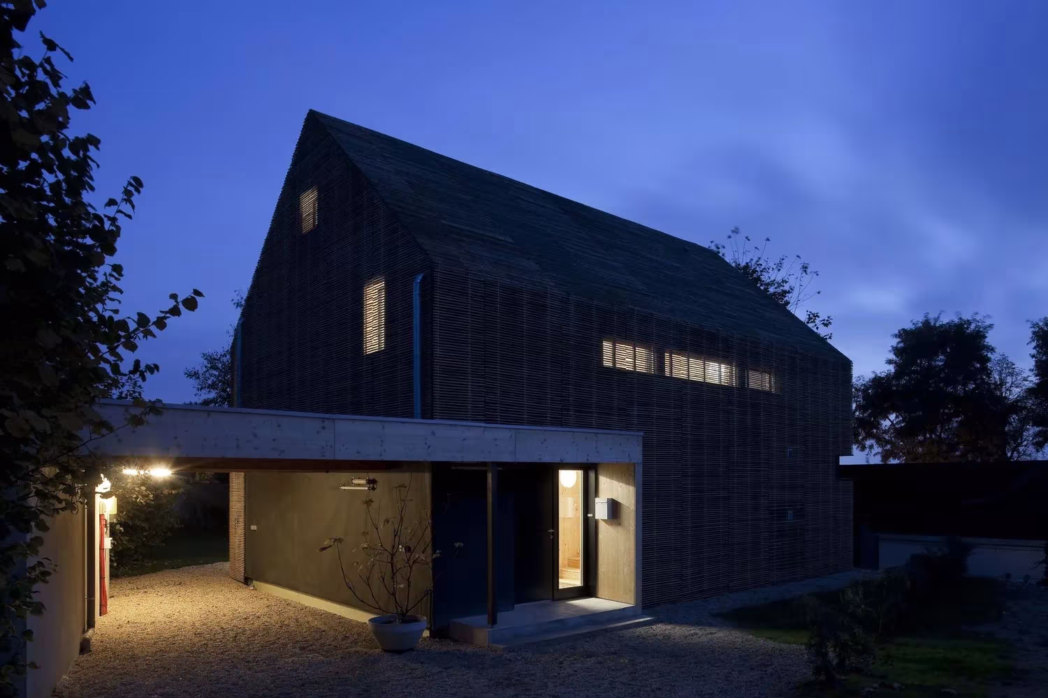 Maison Bambou al anochecer, con su volumen oscuro a dos aguas y el acceso cubierto iluminado, en Bessancourt
