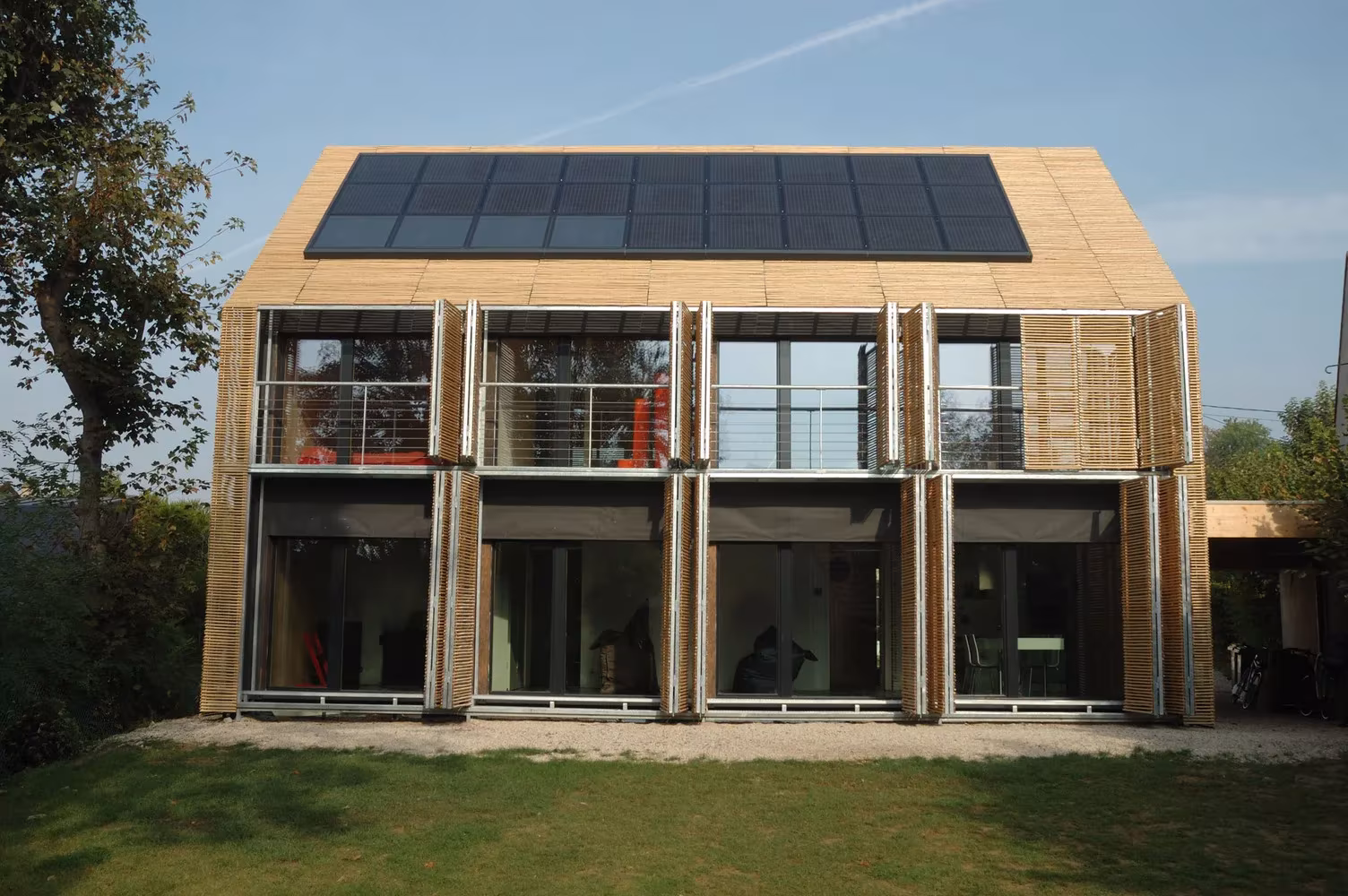 Fachada sur de la Maison Bambou con paneles solares en cubierta, balcones y contraventanas de bambú, Bessancourt