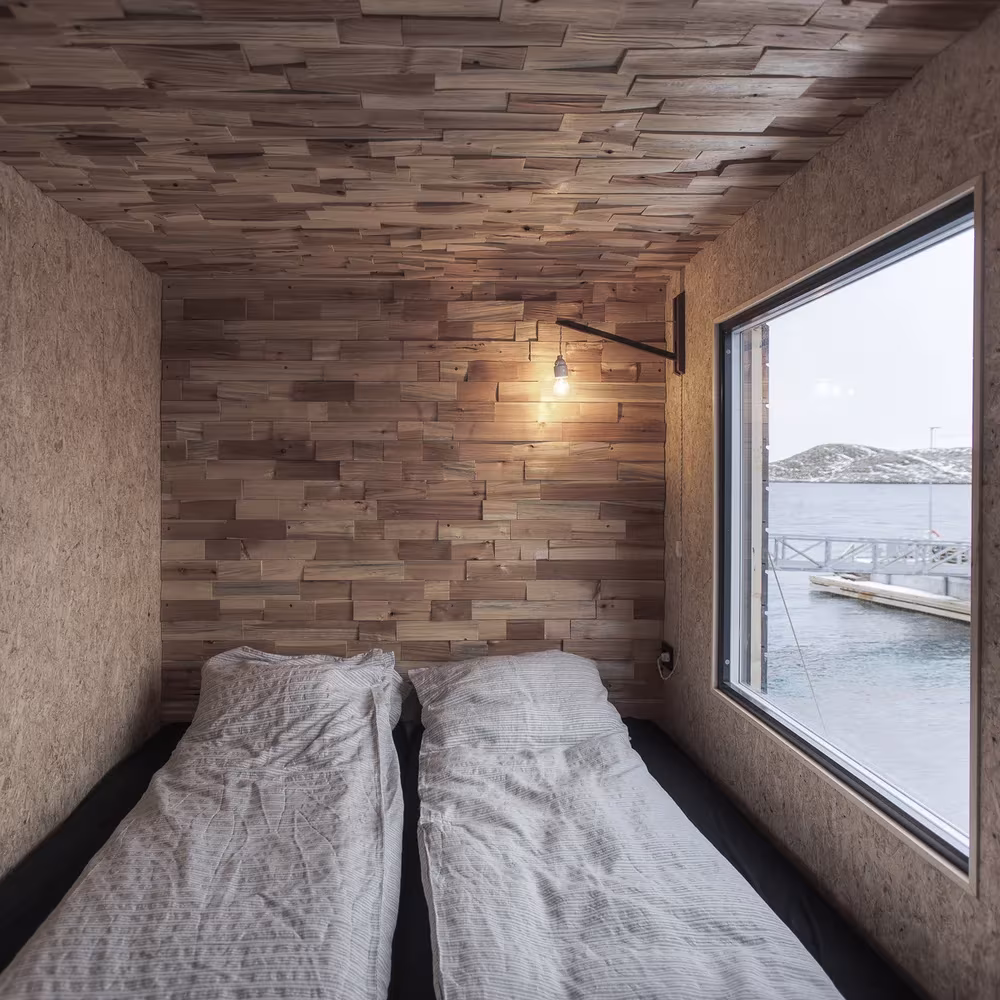 Dormitorio revestido de tablas de madera con dos camas y ventana al fiordo en el refugio de Fleinvær, Noruega