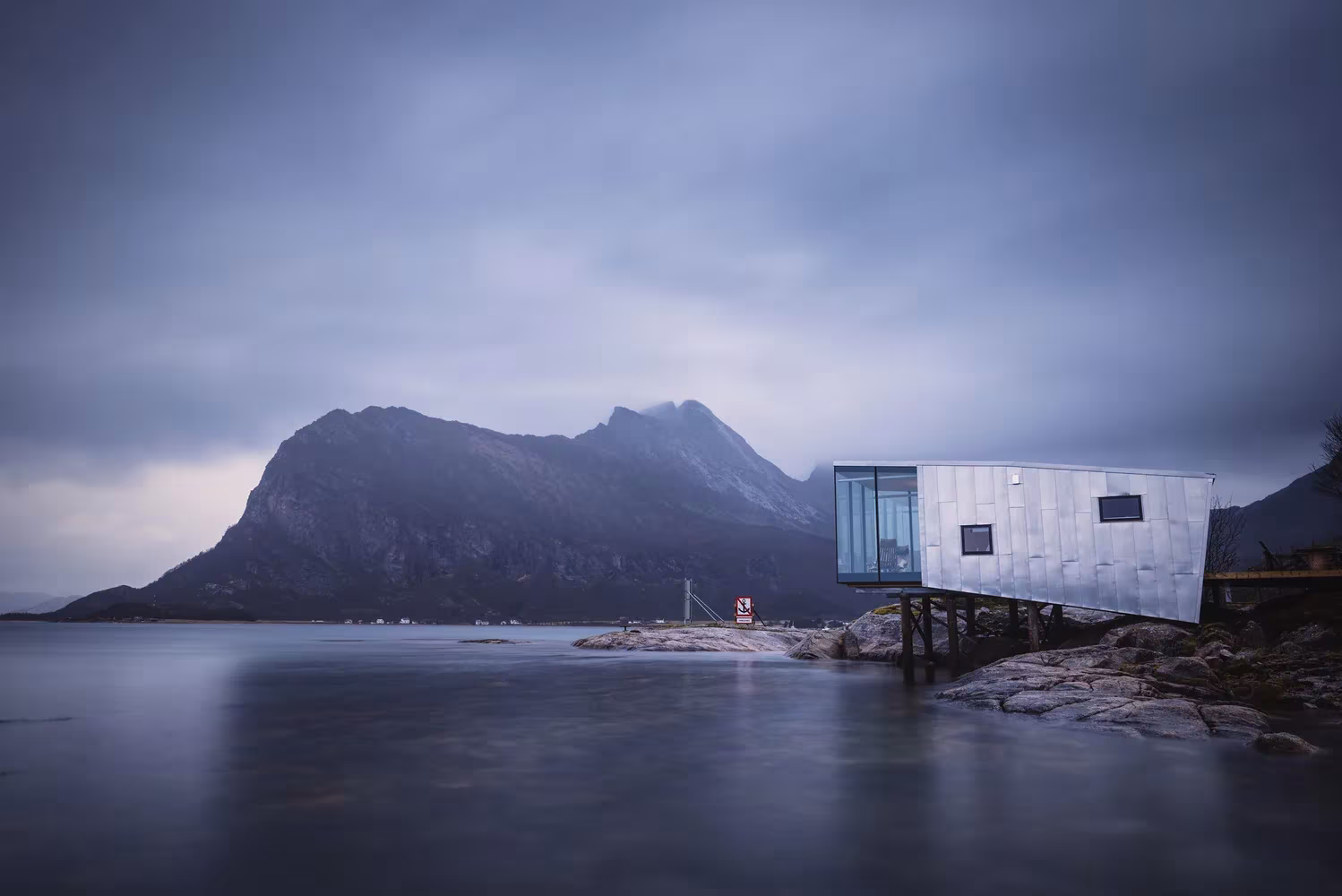 Cabaña de Manshausen volada sobre pilotes frente al fiordo y las montañas bajo un cielo plomizo, Noruega