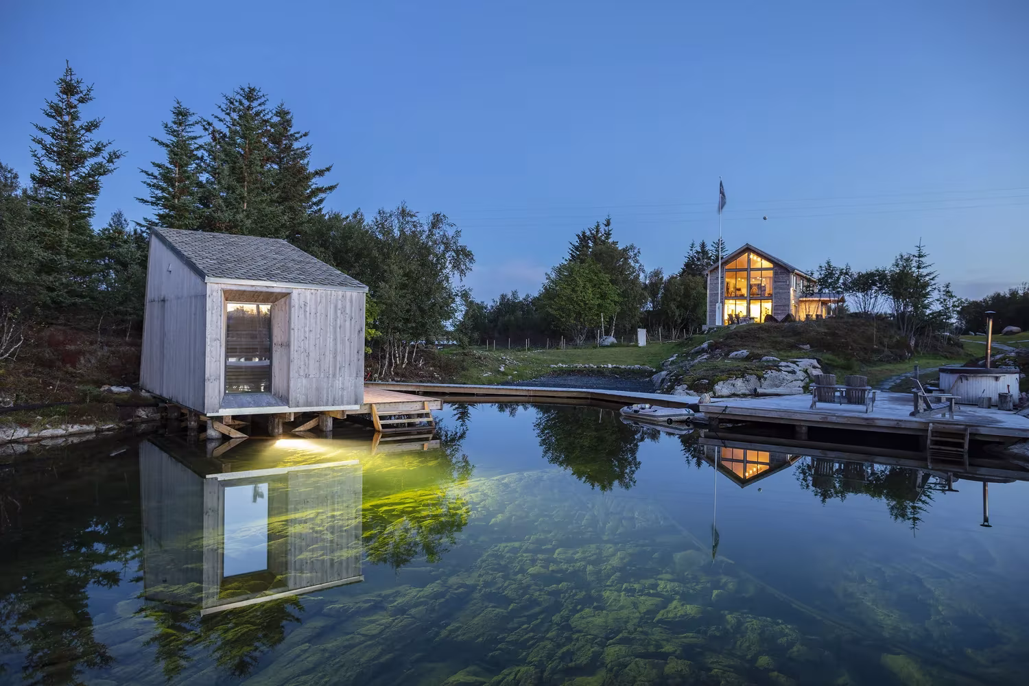 Cabaña de madera junto al agua tranquila y el embarcadero de Manshausen, de Stinessen Arkitektur, al atardecer
