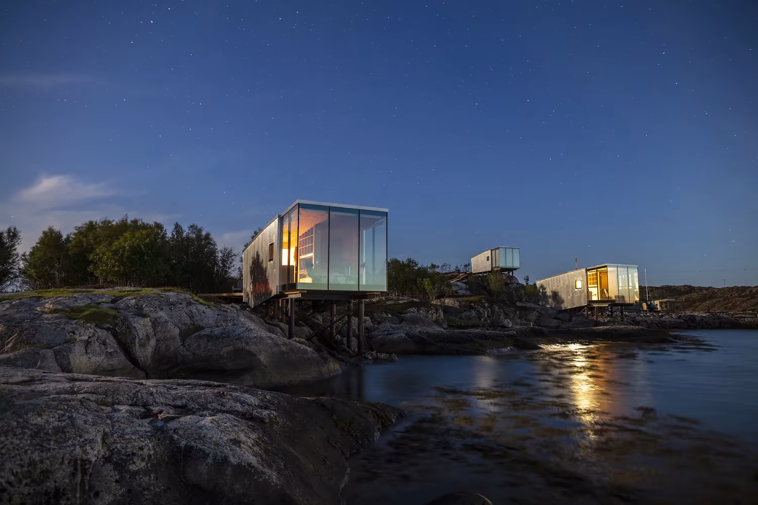 Cabañas de Manshausen iluminadas al anochecer sobre la roca y el fiordo, de Stinessen Arkitektur, Noruega