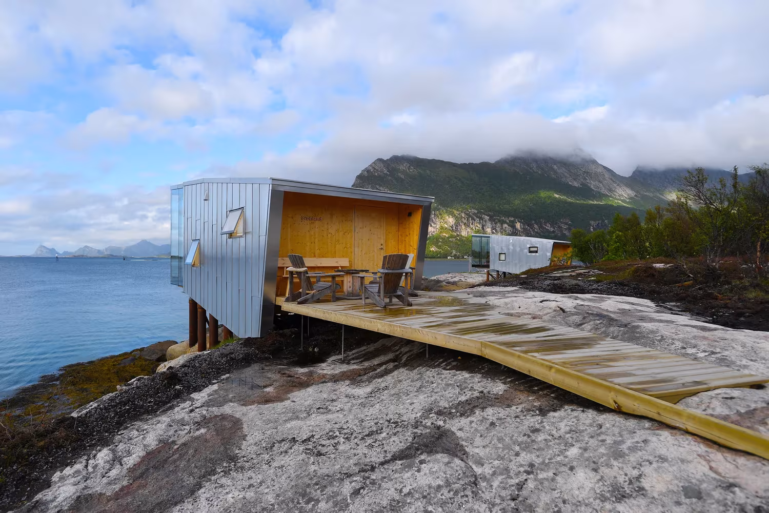 Cabaña de Manshausen con terraza de madera amarilla sobre la roca y vistas al mar y a las montañas, Noruega