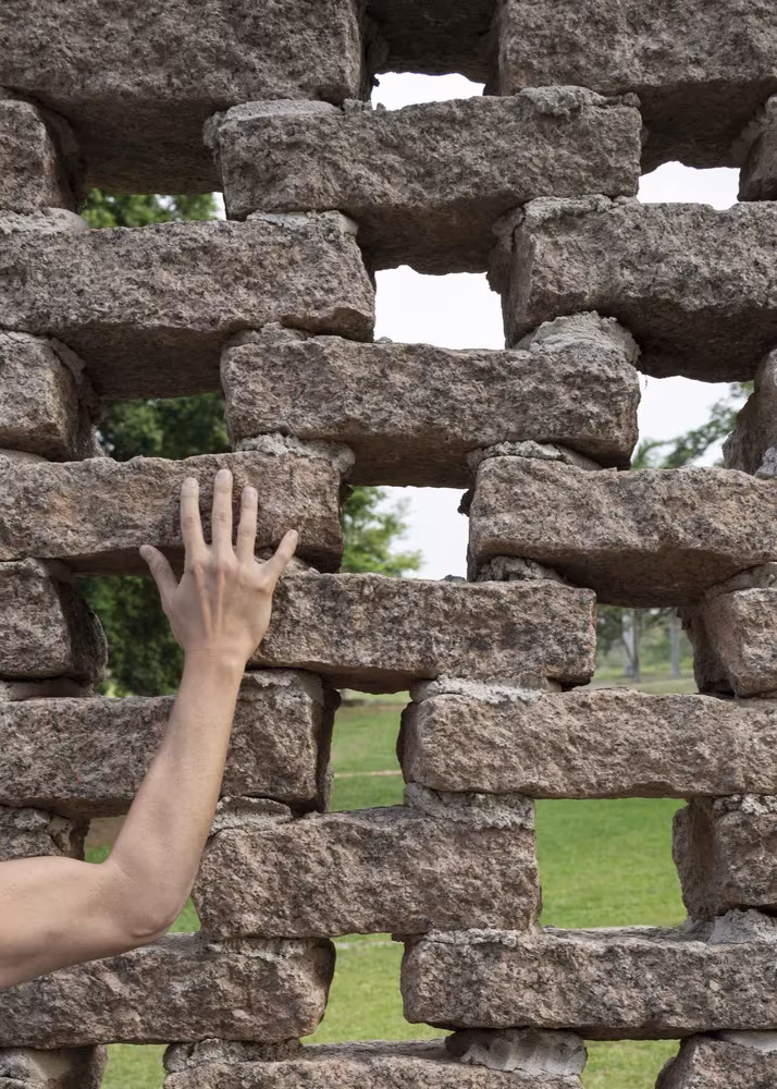 Mano apoyada en el muro de ladrillo calado de la Capilla Ingá-mirim, con el jardín visible entre las piezas