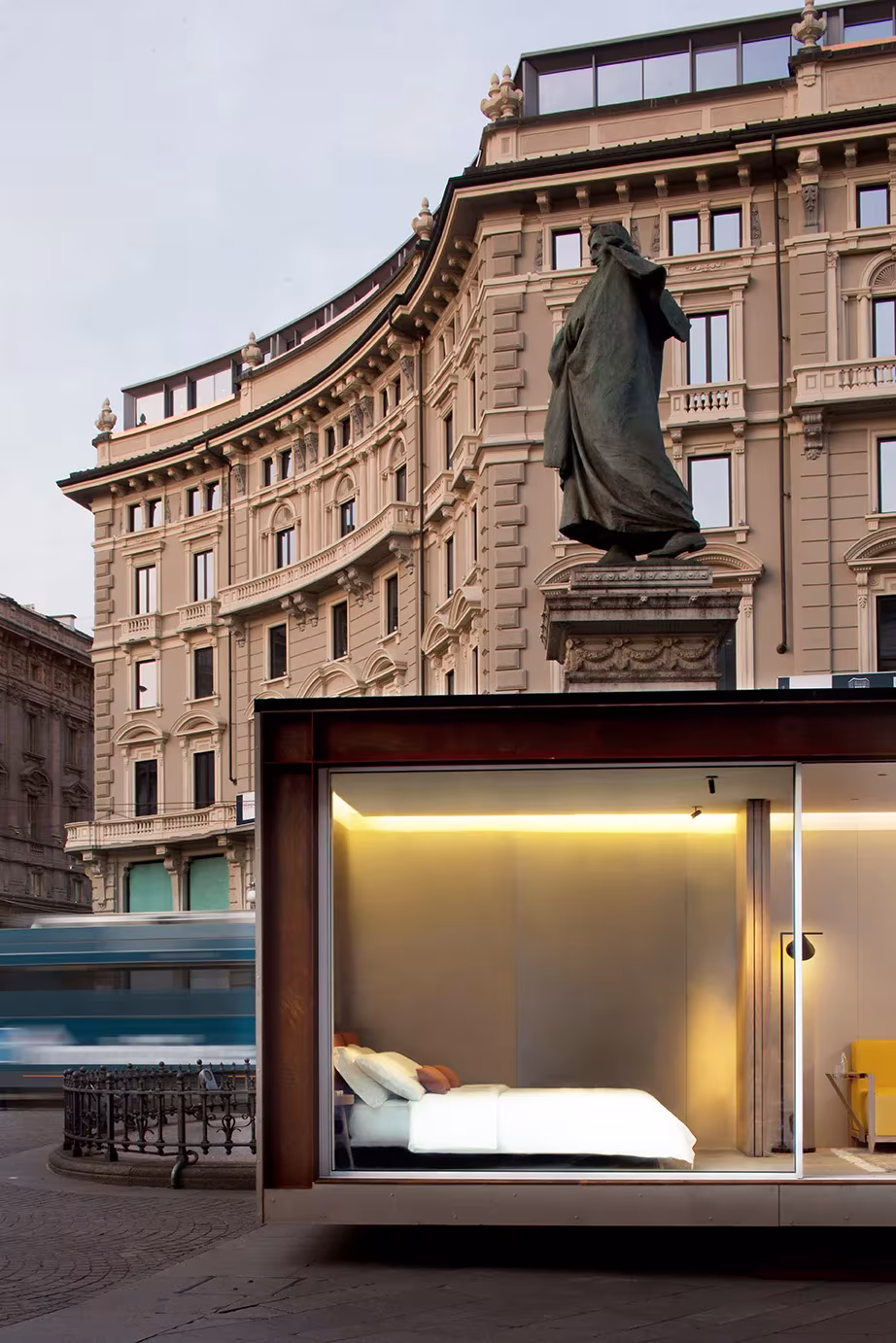 Módulo NO.MADE de acero corten con cama y luz cálida ante un edificio histórico y una estatua, en una plaza de Italia