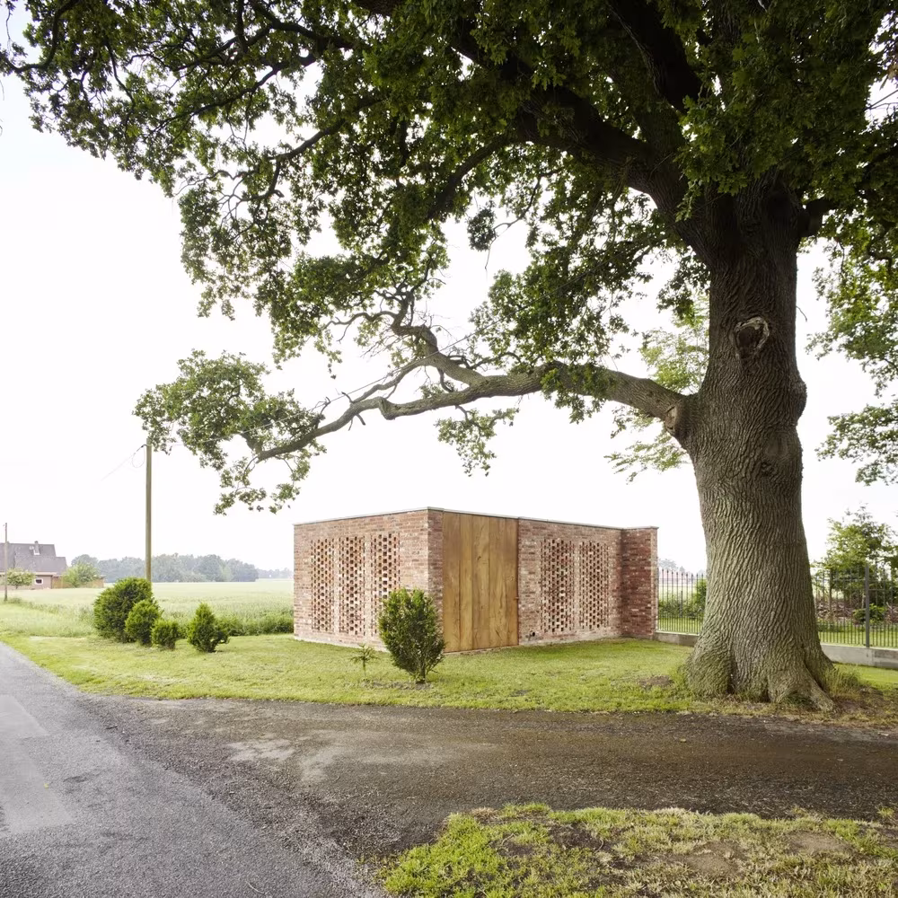 Remisenpavillon de Wirth Architekten, cubo de ladrillo calado y madera junto a un gran roble en un camino rural alemán