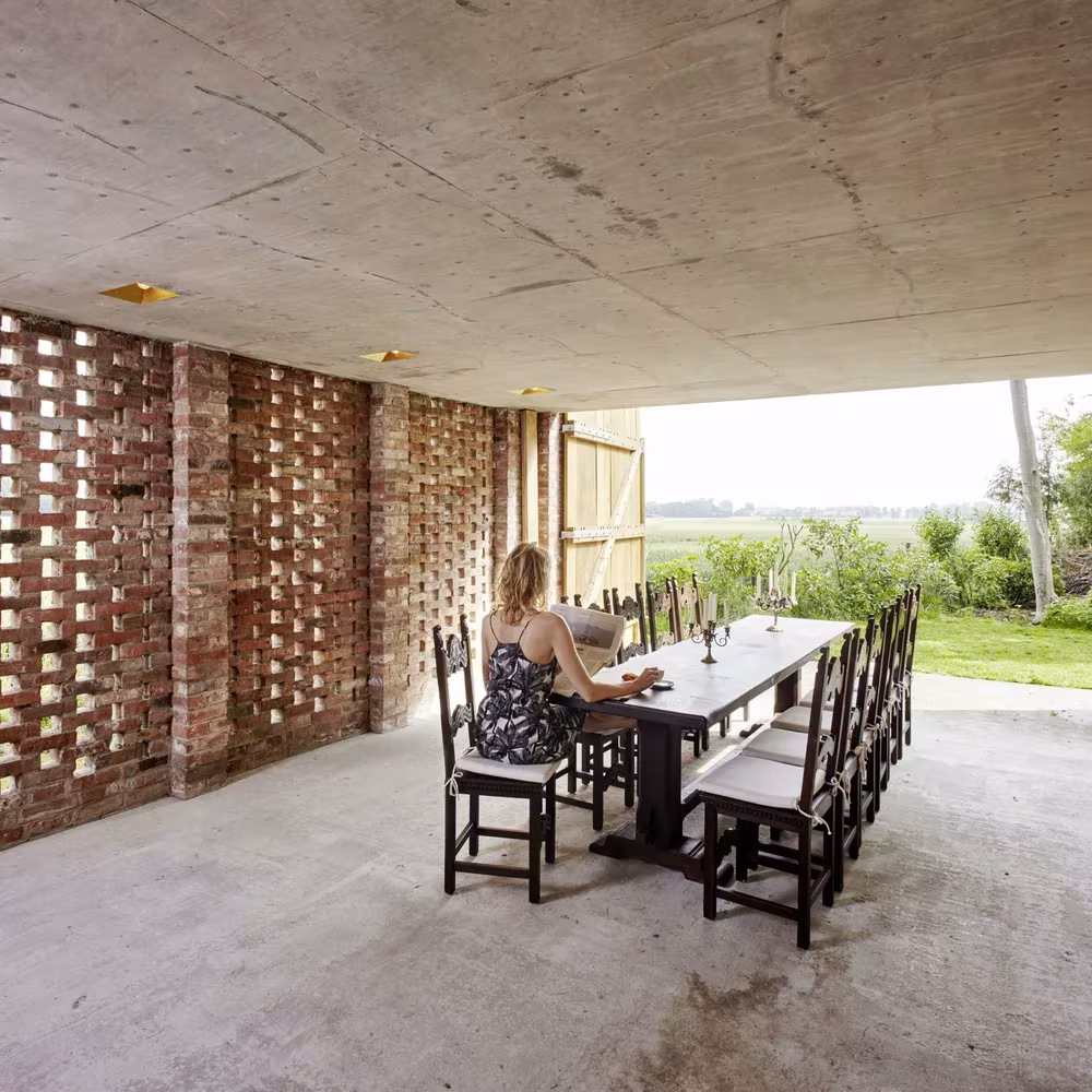 Mesa larga de comedor con sillas de madera en el Remisenpavillon, bajo un techo de hormigón y entre muros de ladrillo