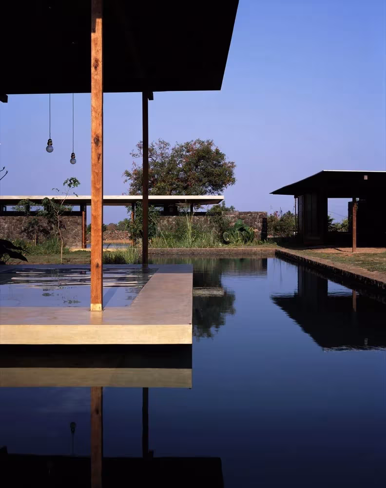 Estanque de Utsav House con una plataforma de hormigón, un pilar de madera y los pabellones al fondo, Maharashtra