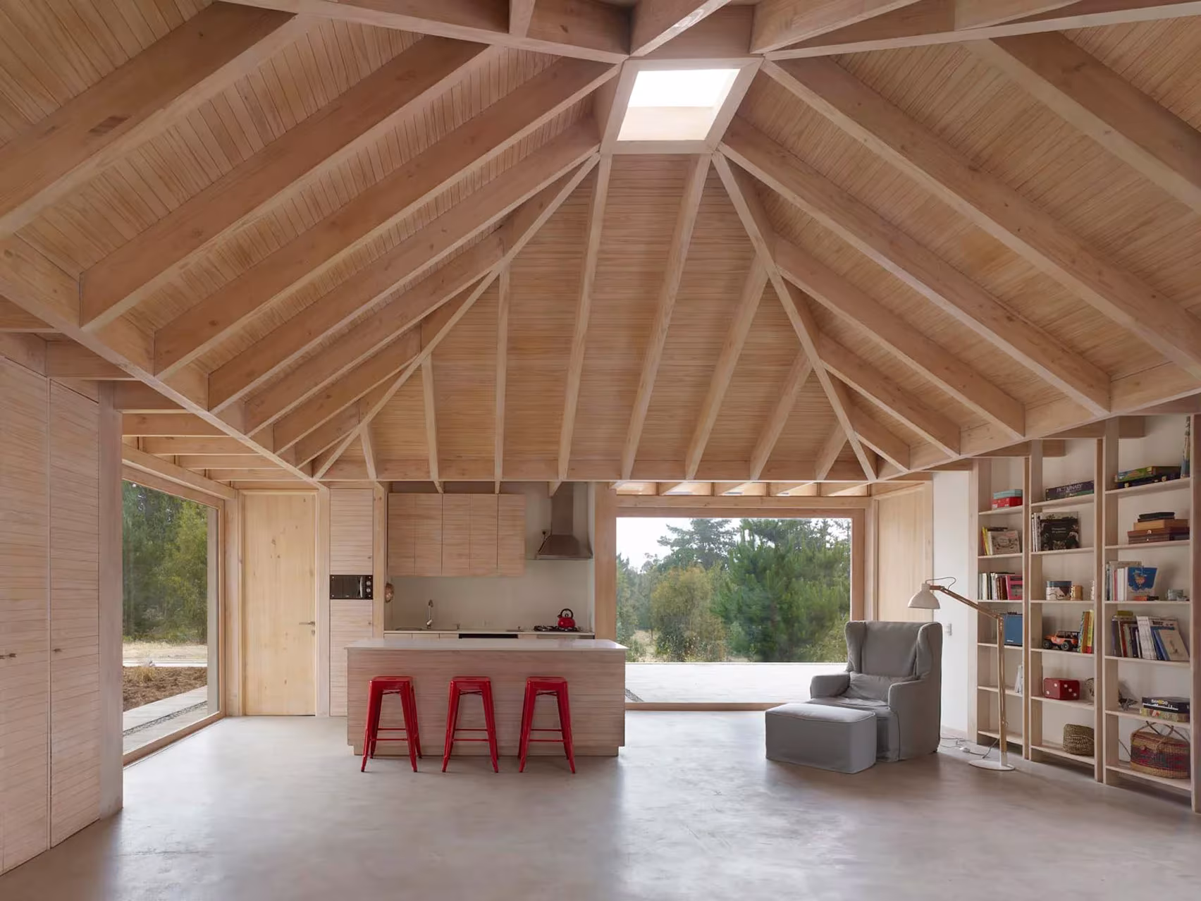 Interior de El Peumo con cubierta de vigas de madera en molinete, cocina con taburetes rojos y librería