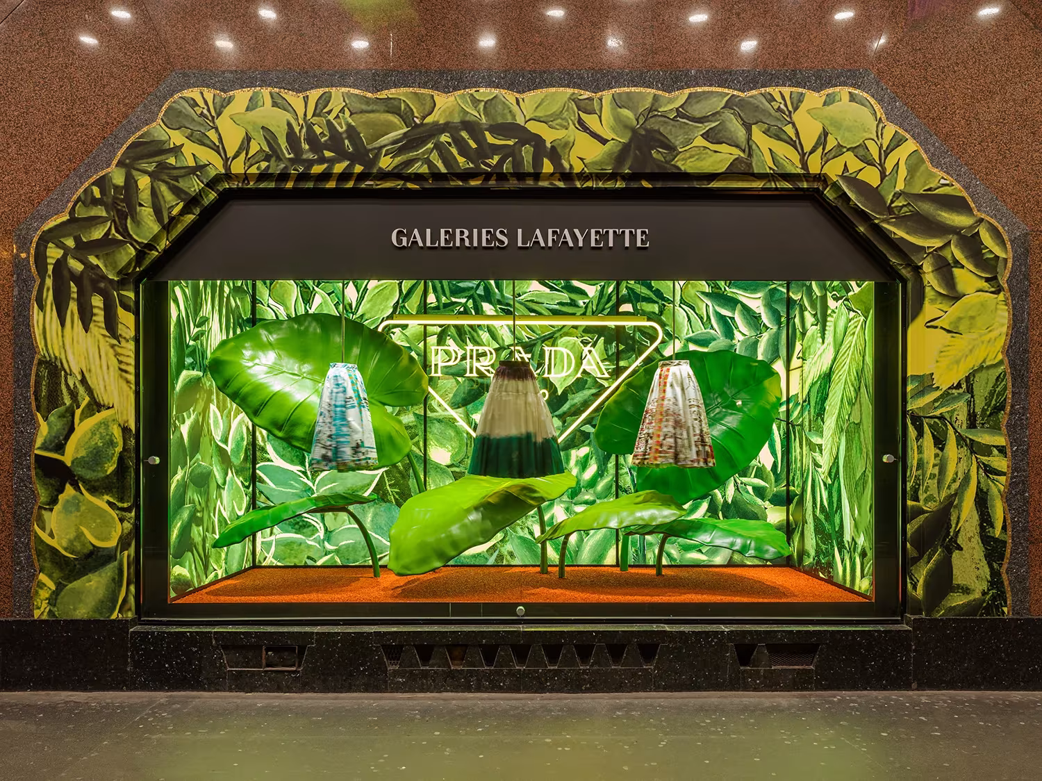 Escaparate de Galeries Lafayette con bolsos de Prada entre hojas verdes sobre un fondo de follaje ilustrado