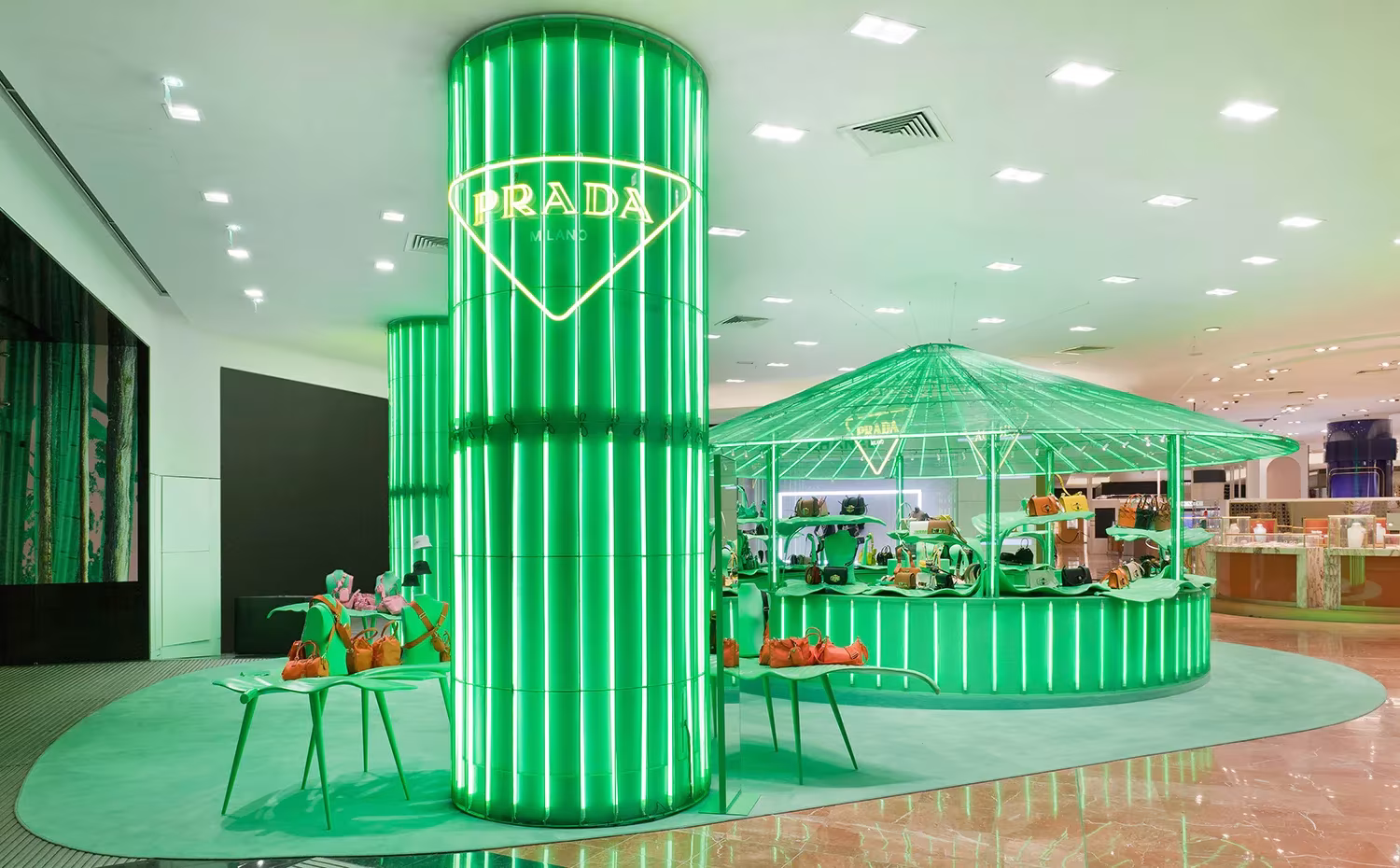 Columna verde luminosa con el logotipo de Prada junto a un pabellón con cubierta de hojas en Galeries Lafayette