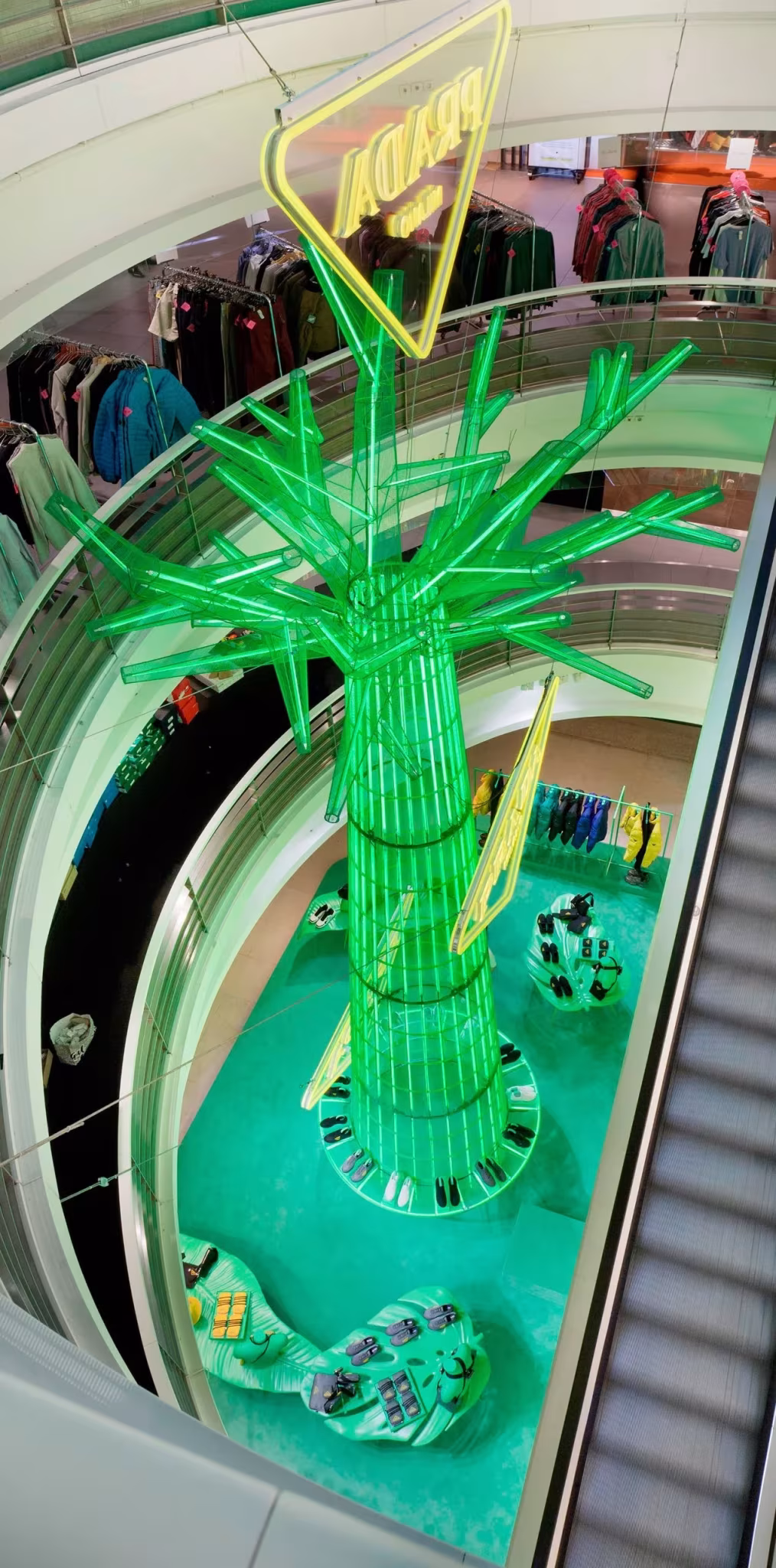 Vista desde arriba de un árbol verde luminoso de Prada que se eleva por el hueco de las escaleras mecánicas