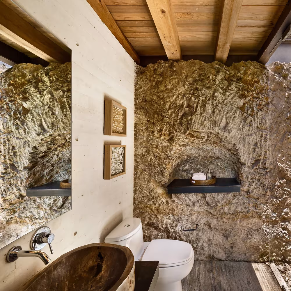 Baño de la Casa Tucán con muro de roca natural, lavabo de madera tallada y techo de vigas de madera