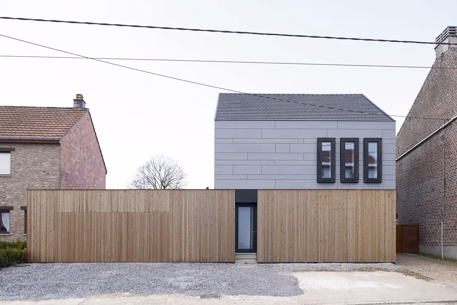 Fachada a la calle de Goetsenhoven House, con muro de lamas de madera y volumen gris con tres ventanas verticales