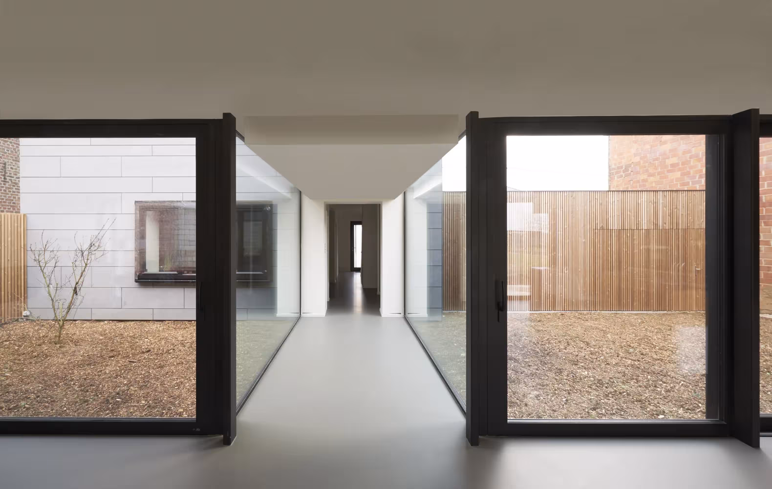 Pasillo acristalado de Goetsenhoven House que une dos patios con grava y fachada de lamas de madera