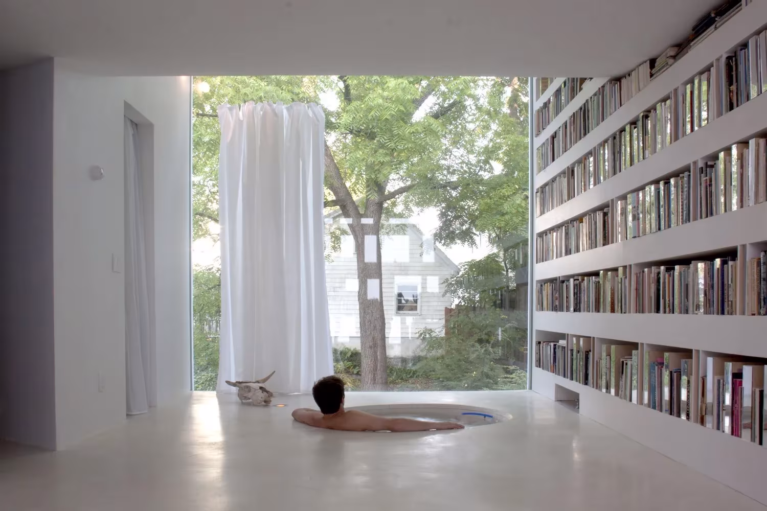 Sala de la biblioteca de Haffenden House con un ventanal a los árboles, cortina blanca y estantes hasta el techo
