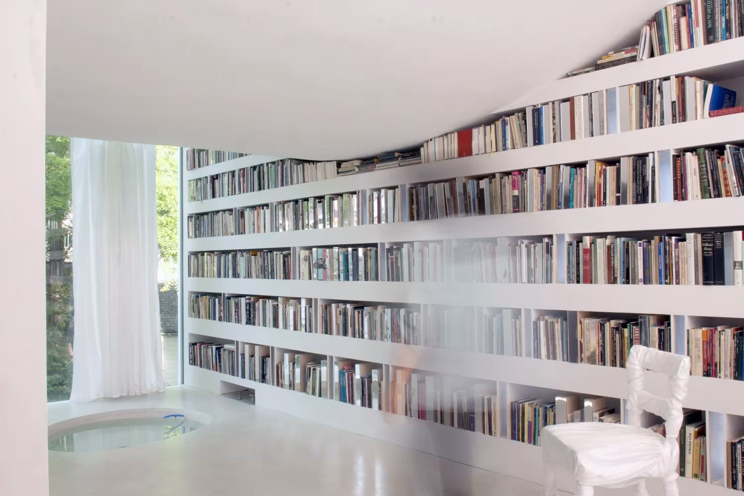 Estantería blanca que recorre el muro curvo de la biblioteca de Haffenden House, con una cortina junto al ventanal