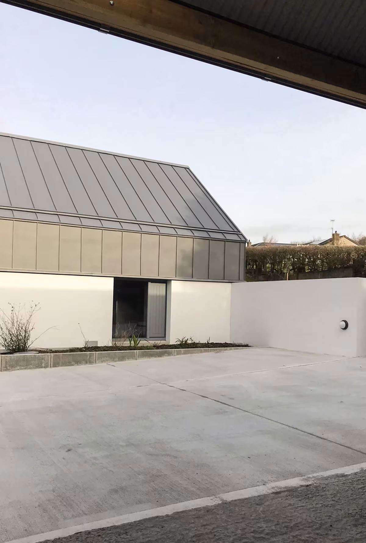 Patio de hormigón ante el muro blanco y el volumen de zinc de House Lessans, con el vuelo de la cubierta en primer plano