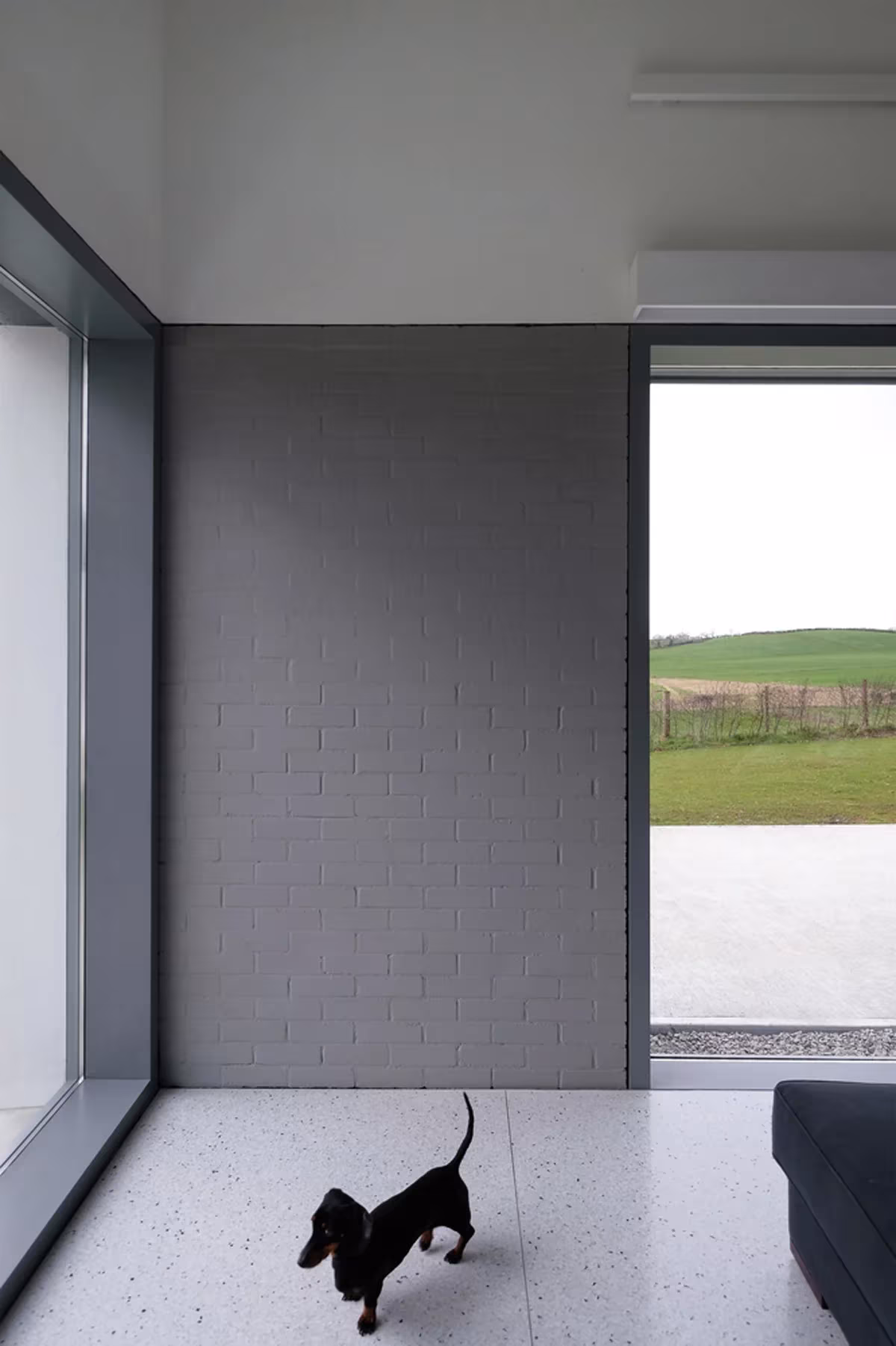 Interior de House Lessans con muro de bloque gris, suelo claro y un gran vano abierto al prado