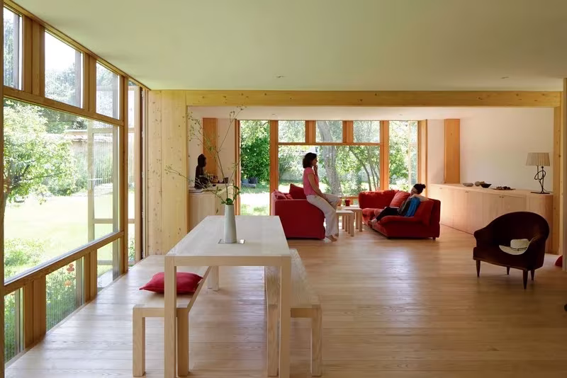 Salón-comedor acristalado con mesa larga de madera y sofás rojos que se abre al jardín en la Maison dans le Jardin