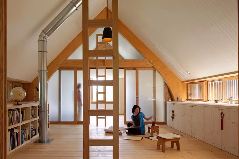 Sala bajo la cubierta a dos aguas con estantería, escalera de madera y paneles translúcidos en la Maison dans le Jardin