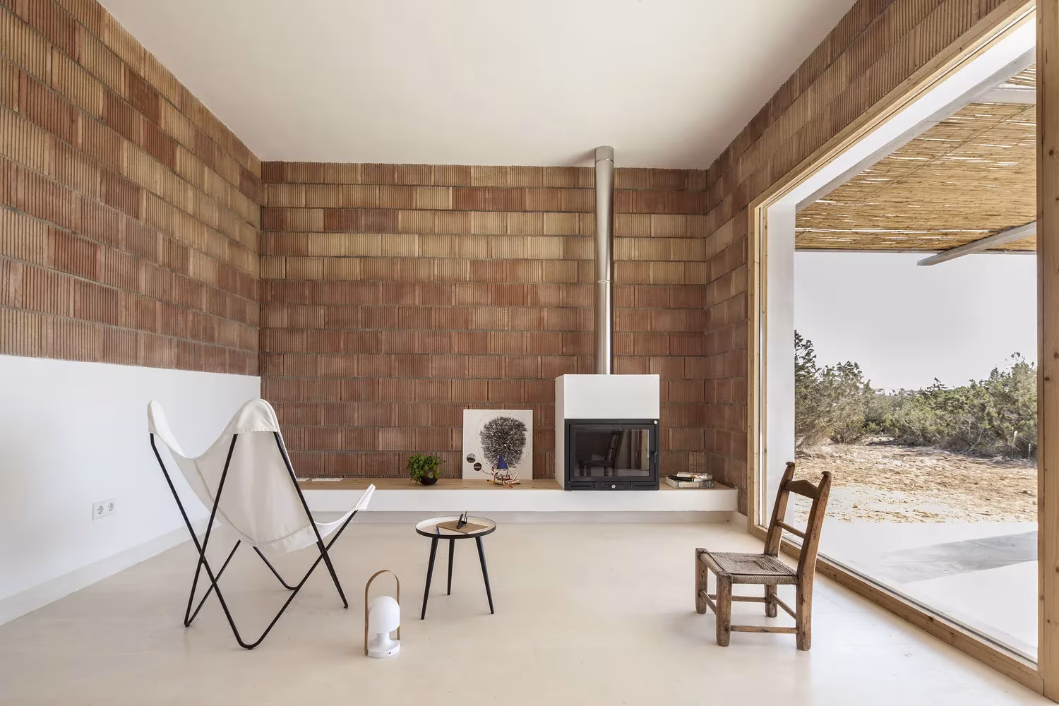 Salón de Can Xomeu Rita con muro de ladrillo cerámico visto, chimenea y gran ventanal con carpintería de madera