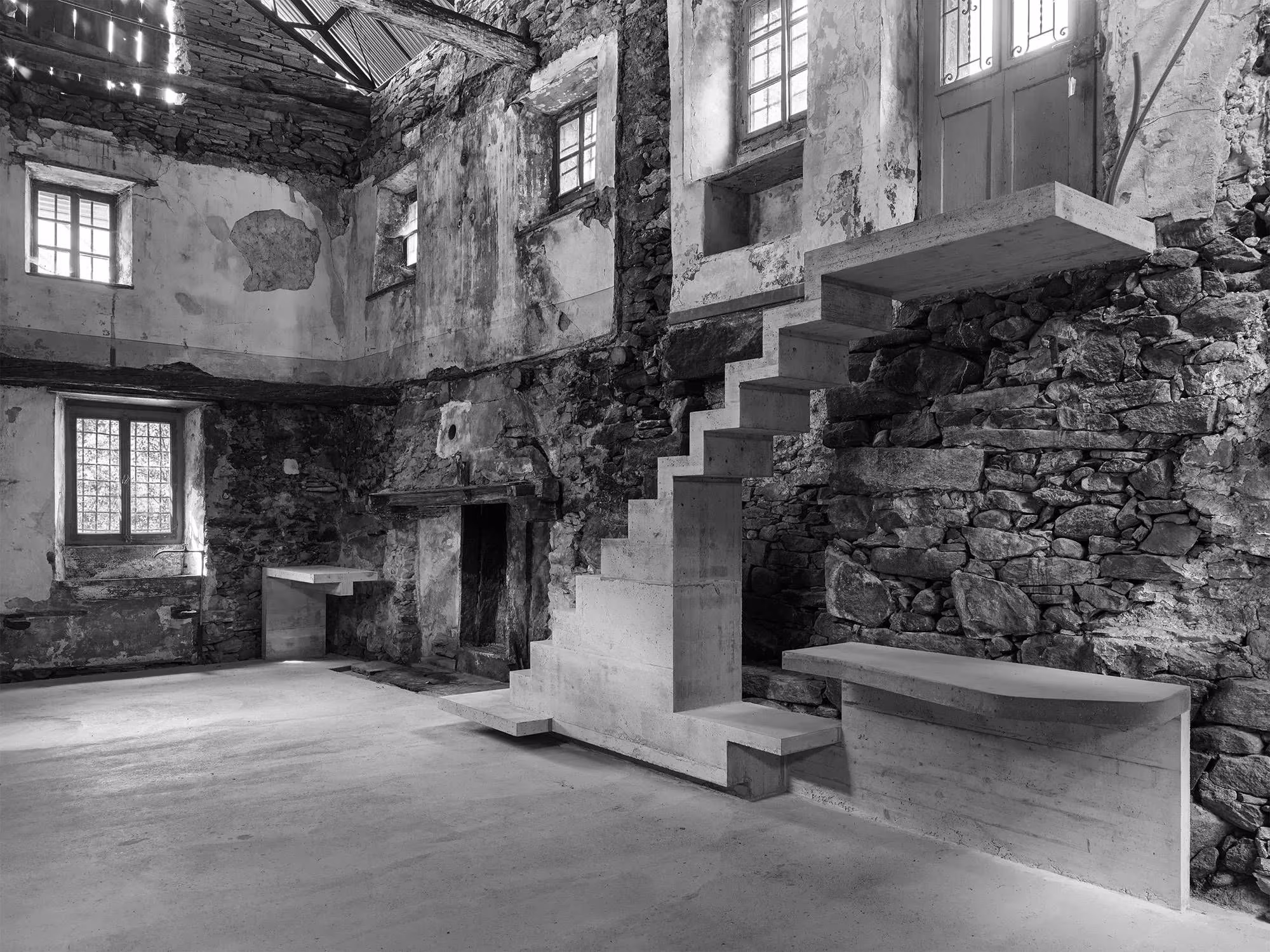 Sala de Casa Mosogno con escalera de hormigón volada, muros de piedra y ventanas con contraventanas