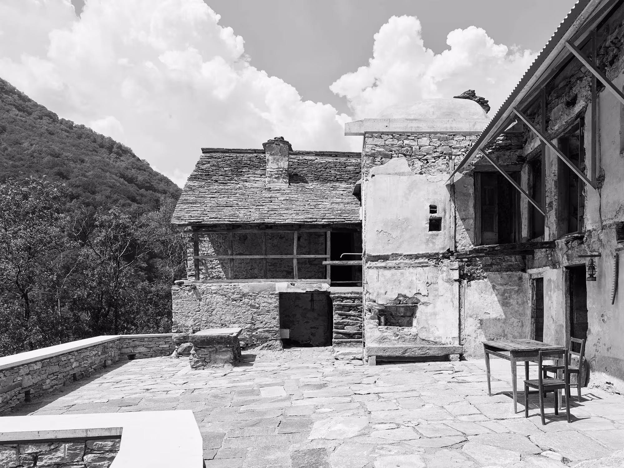 Terraza de losas ante las construcciones de piedra de Casa Mosogno, con una mesa y sillas al aire libre, en blanco y negro
