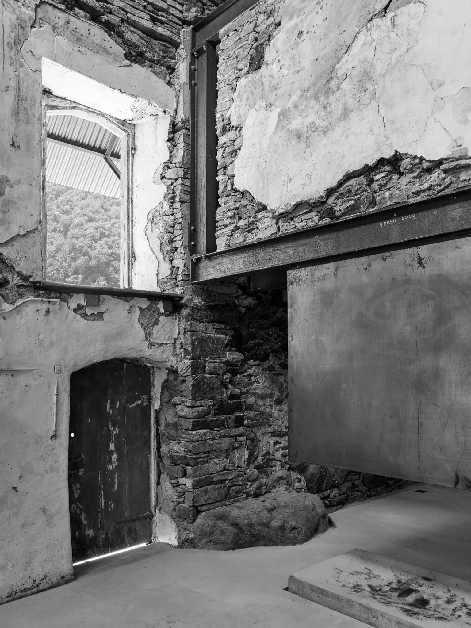 Vano de piedra y viga de acero en el interior de Casa Mosogno, con una puerta de madera y paredes desconchadas