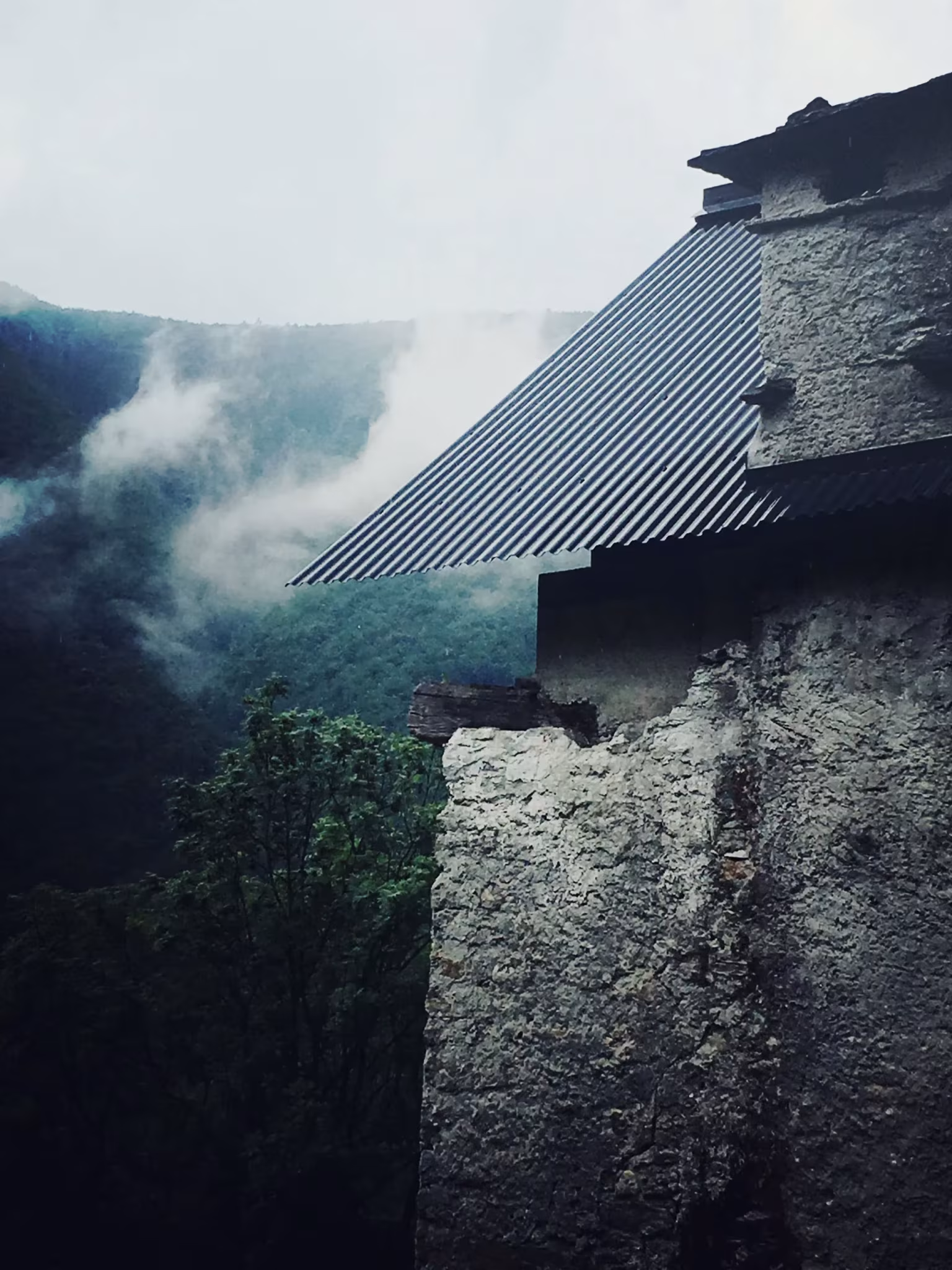 Esquina de piedra de Casa Mosogno con la cubierta de chapa metálica inclinada y el valle entre nubes al fondo
